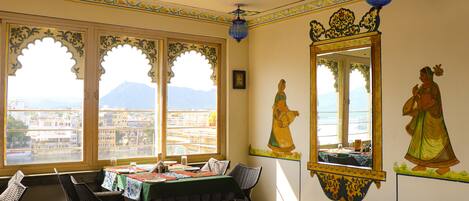 A dining area with a table set for two, a large mirror, and a view of a cityscape through arched windows.