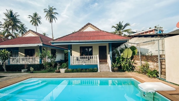 Ein Swimmingpool mit Liegestühlen, ein Haus mit rotem Dach und Palmen im Hintergrund.
