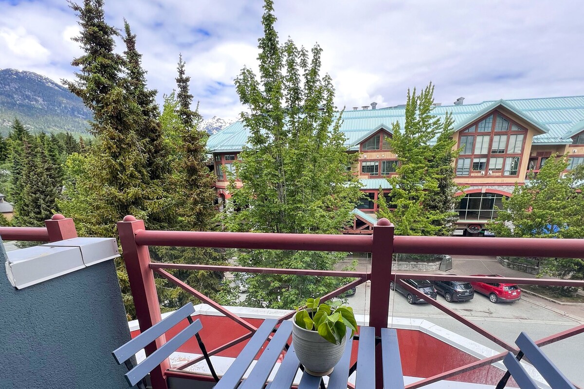 A balcony with a potted plant, overlooking a parking area and buildings with green roofs.