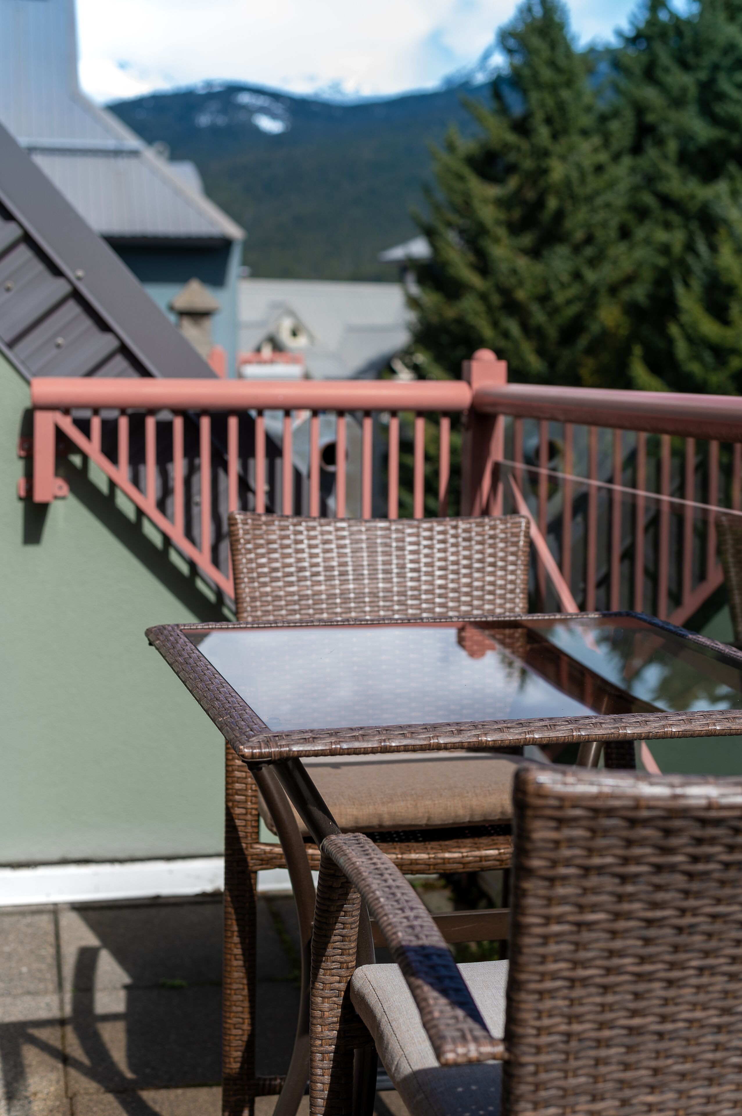 A wicker table and chairs on a balcony with a red railing, overlooking a mountain and greenery.