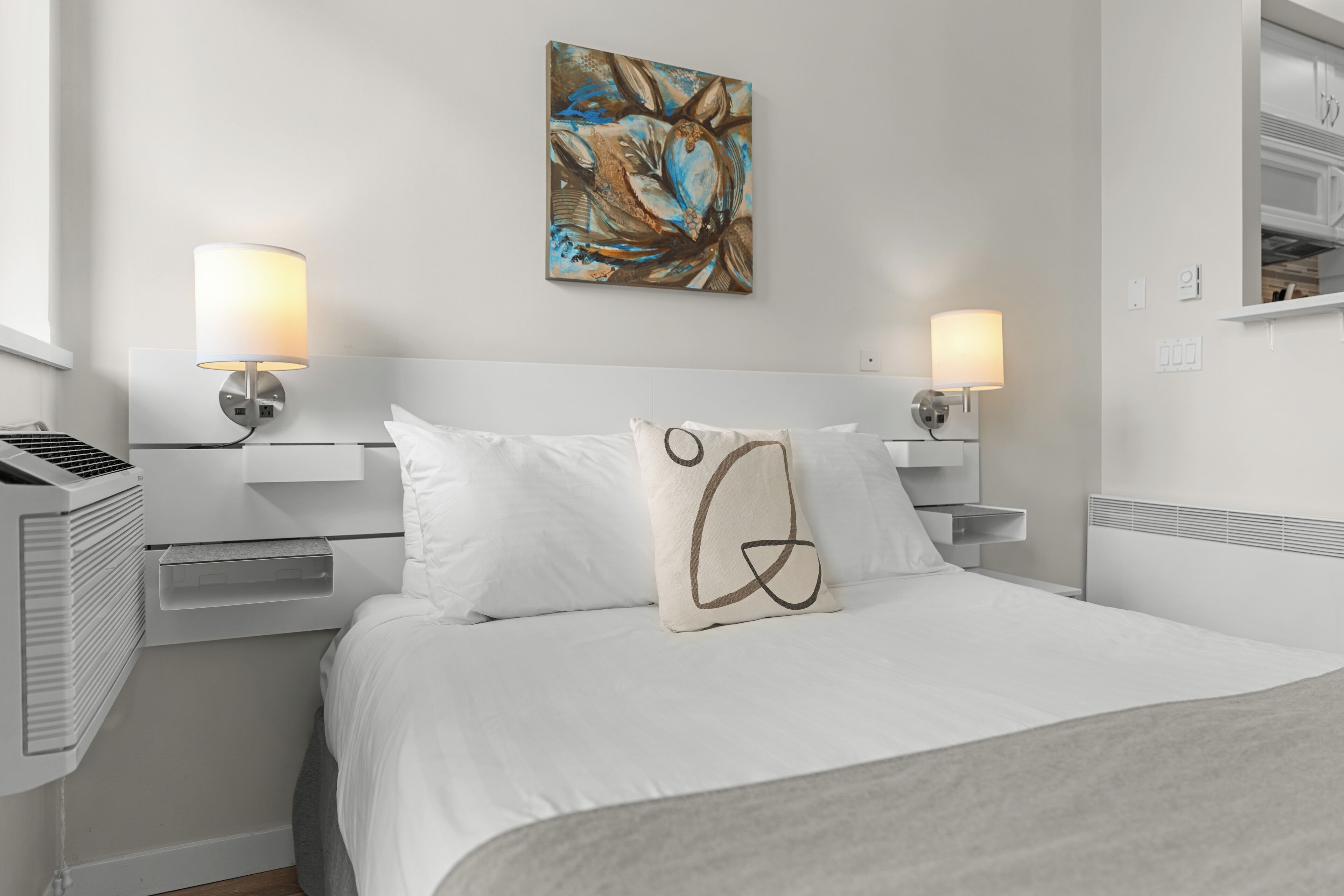 A hotel room with a bed, two bedside lamps, a wall-mounted artwork, and a heater.