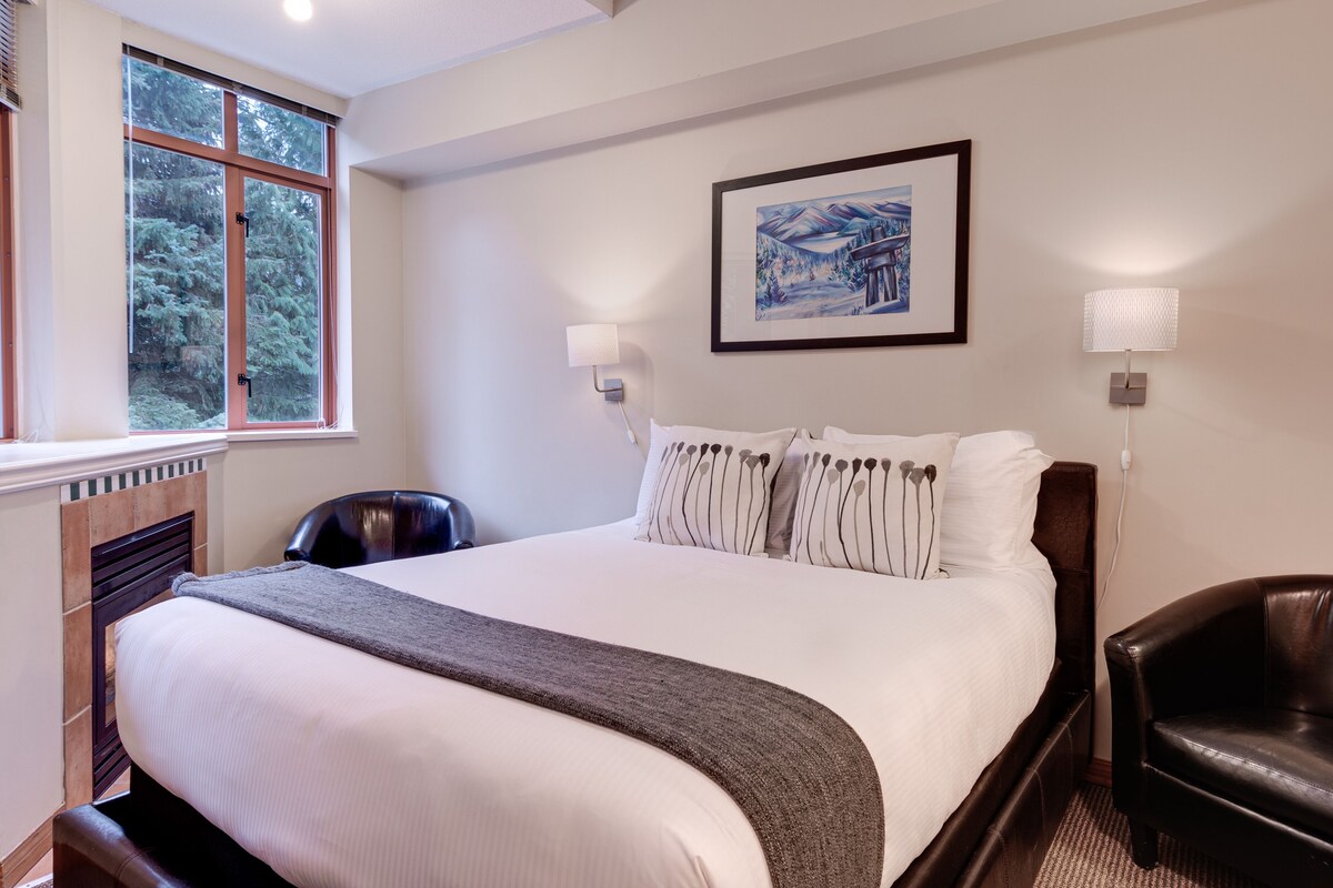 Habitación de hotel con una cama grande, una ventana con vista a árboles, un cuadro enmarcado en la pared y una lámpara en la mesita de noche.