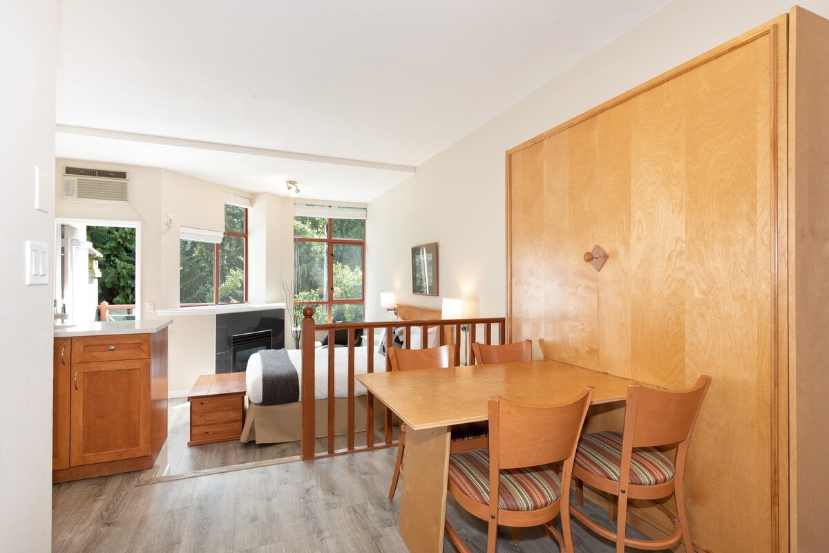 A modern kitchen with wooden cabinets, a dining table with chairs, and a view of the outdoors through large windows.