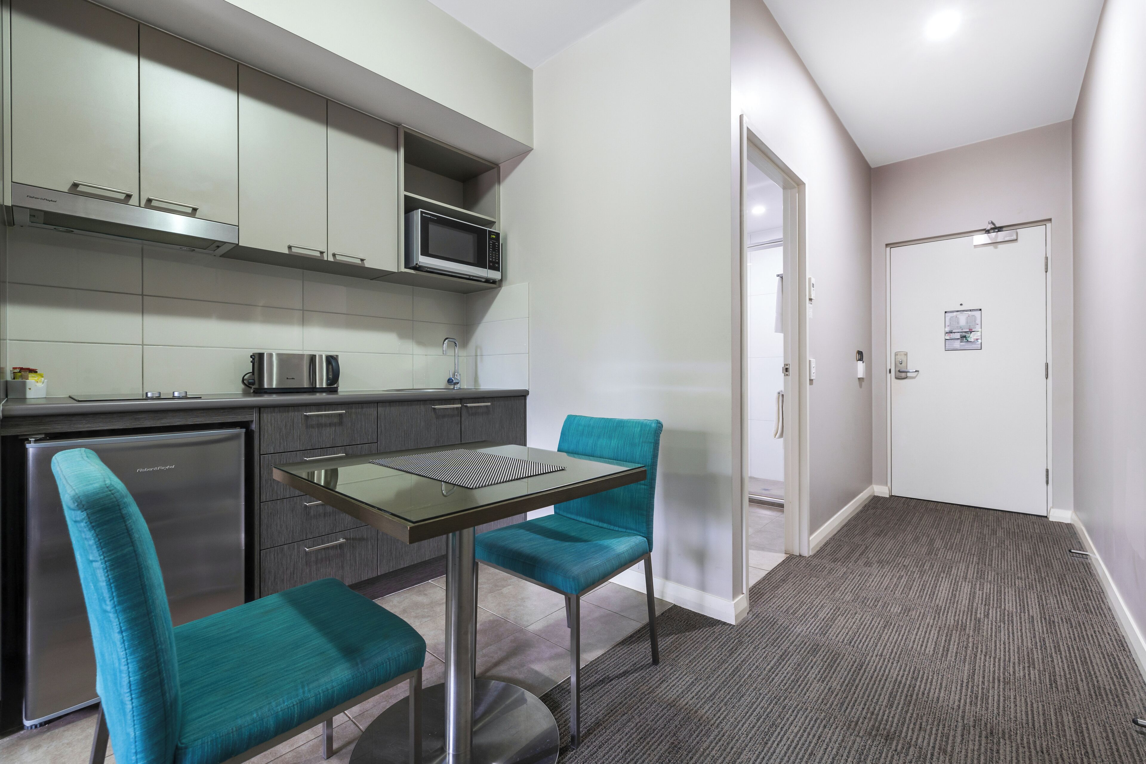 A modern kitchen with a microwave, stainless steel refrigerator, and a glass-top table with teal chairs.