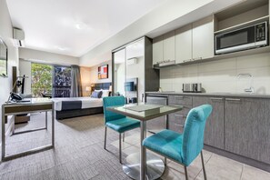A modern hotel room with a glass dining table, teal chairs, a bed with a view, a kitchenette, and a large window. - Hudson Berrimah (Berrimah)