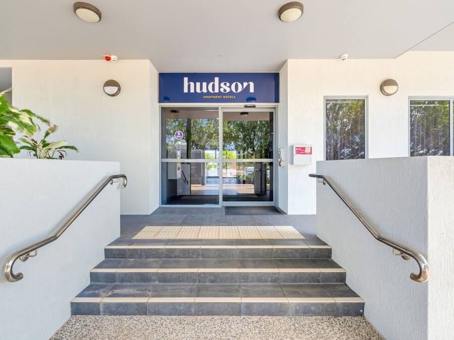 Entrada de un hotel con un letrero que dice "Hudson Apartment Hotels", un conjunto de escaleras con barandillas y una vista del interior a través de las puertas de vidrio.