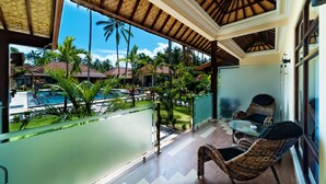 A balcony with wicker chairs and a glass table, overlooking a swimming pool and lush greenery.