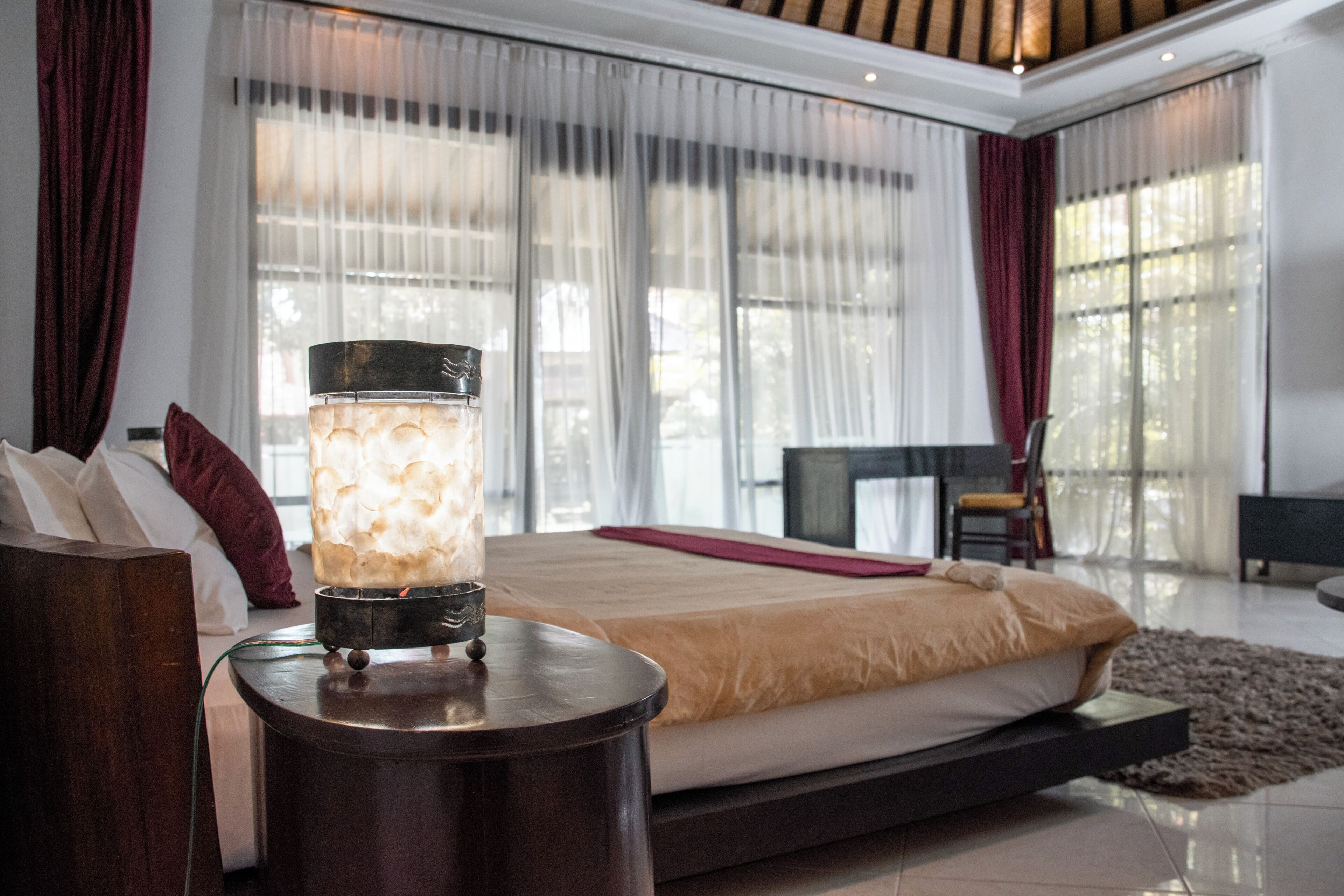 A modern bedroom with a large bed, a bedside table with a lamp, and a view of the outdoors through sheer curtains.