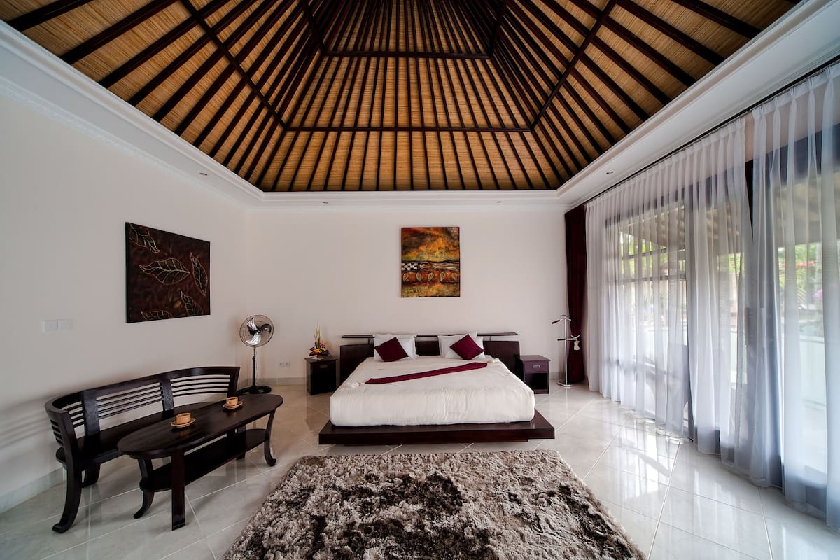 A bedroom with a large bed, a wooden ceiling, a large window with sheer curtains, a dark bench, and a rug.