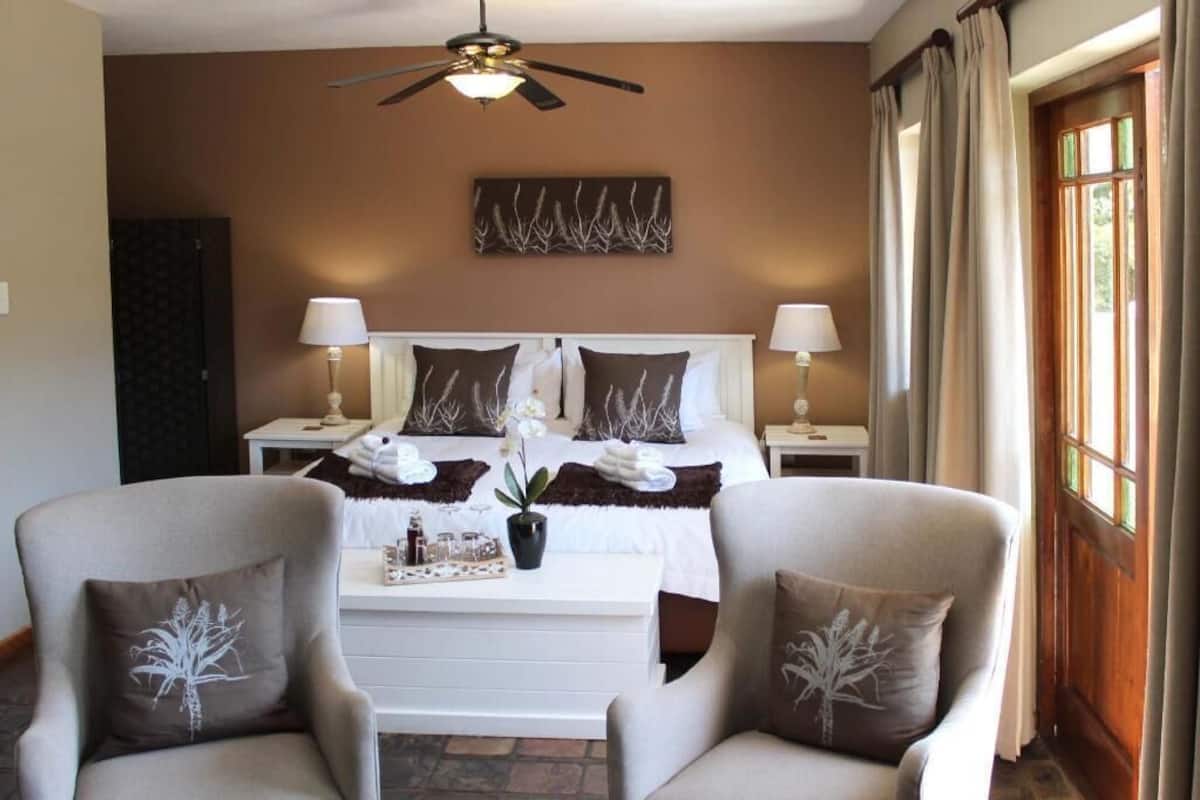 A hotel room with a bed, two bedside tables with lamps, a ceiling fan, a window with curtains, and two armchairs with decorative pillows.