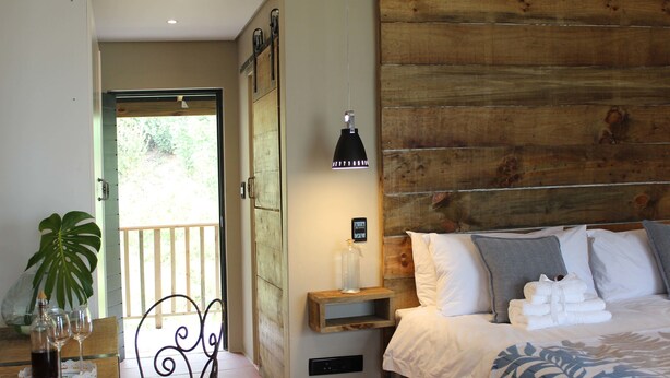 Un dormitorio con cabecera de madera, una mesita de noche con lámpara, una mesita auxiliar y una cama con ropa blanca, con vista al exterior a través de una puerta.
