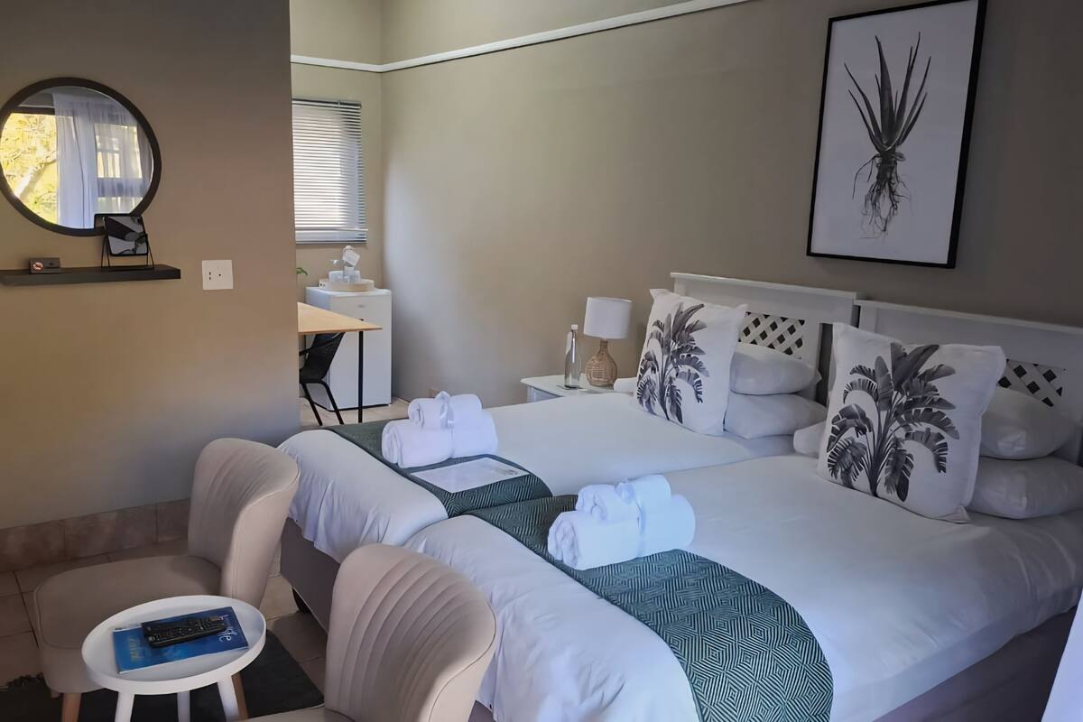 A hotel room with a bed, a small table with a remote control, a chair, a desk with a lamp, and a window with blinds.