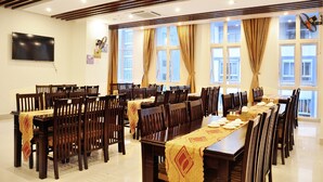 Un salón de conferencias con mesas y sillas largas, ventanas amplias con cortinas y un televisor montado.