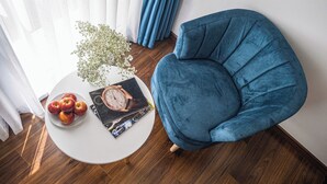 Una habitación con un sillón azul, una mesita blanca con manzanas y una revista, y una ventana con cortinas.