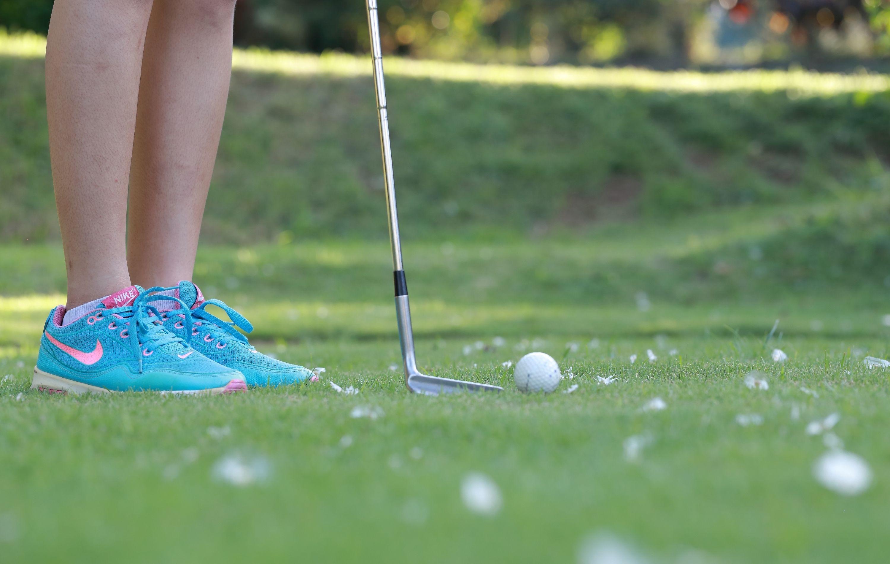 A close-up of a golf club and a golf ball on a green, with a pair of blue and pink shoes.