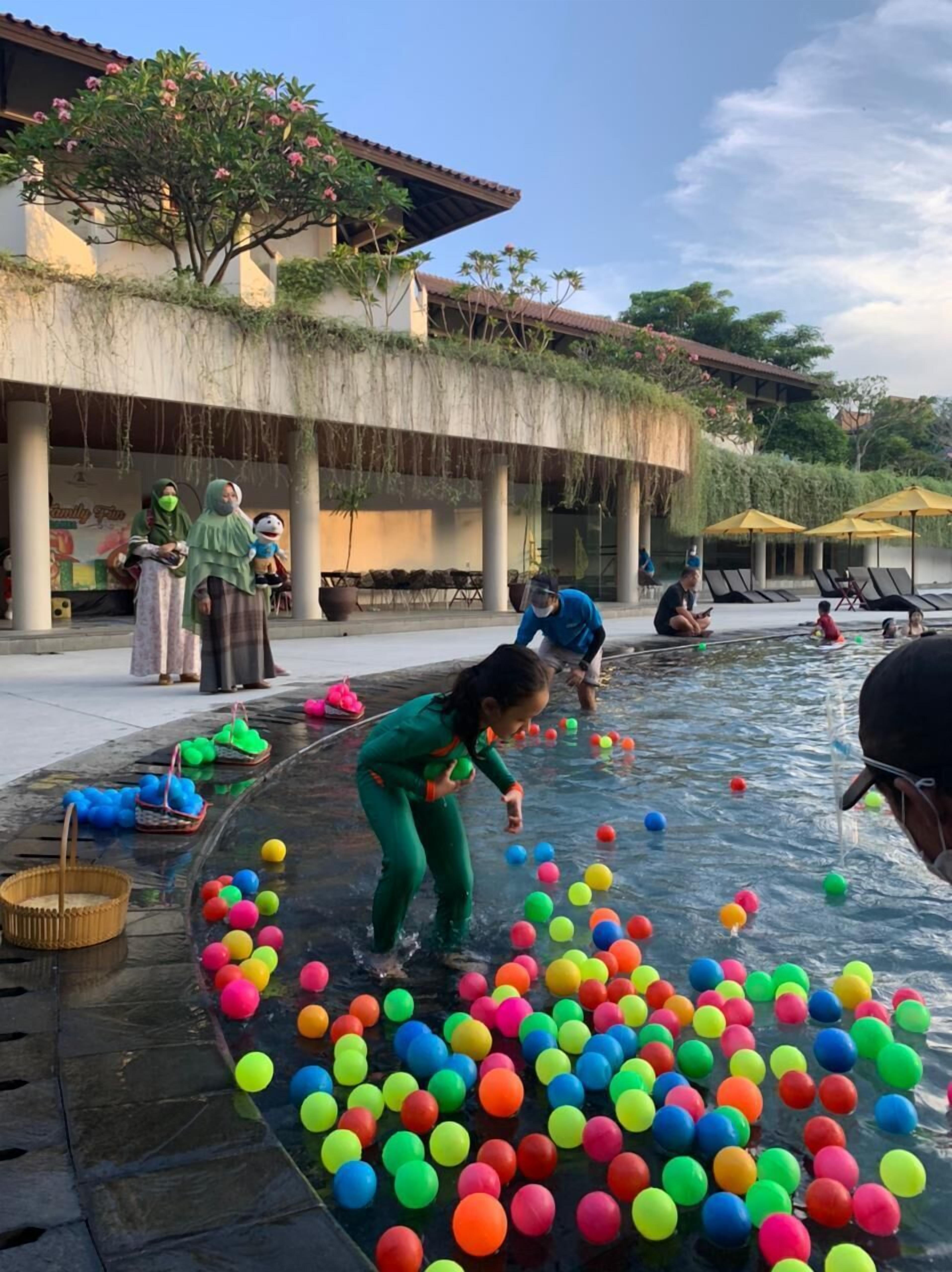 Un espace piscine agrémenté de balles colorées, de chaises longues et d’un bâtiment doté de colonnes et d’un toit en tuiles.