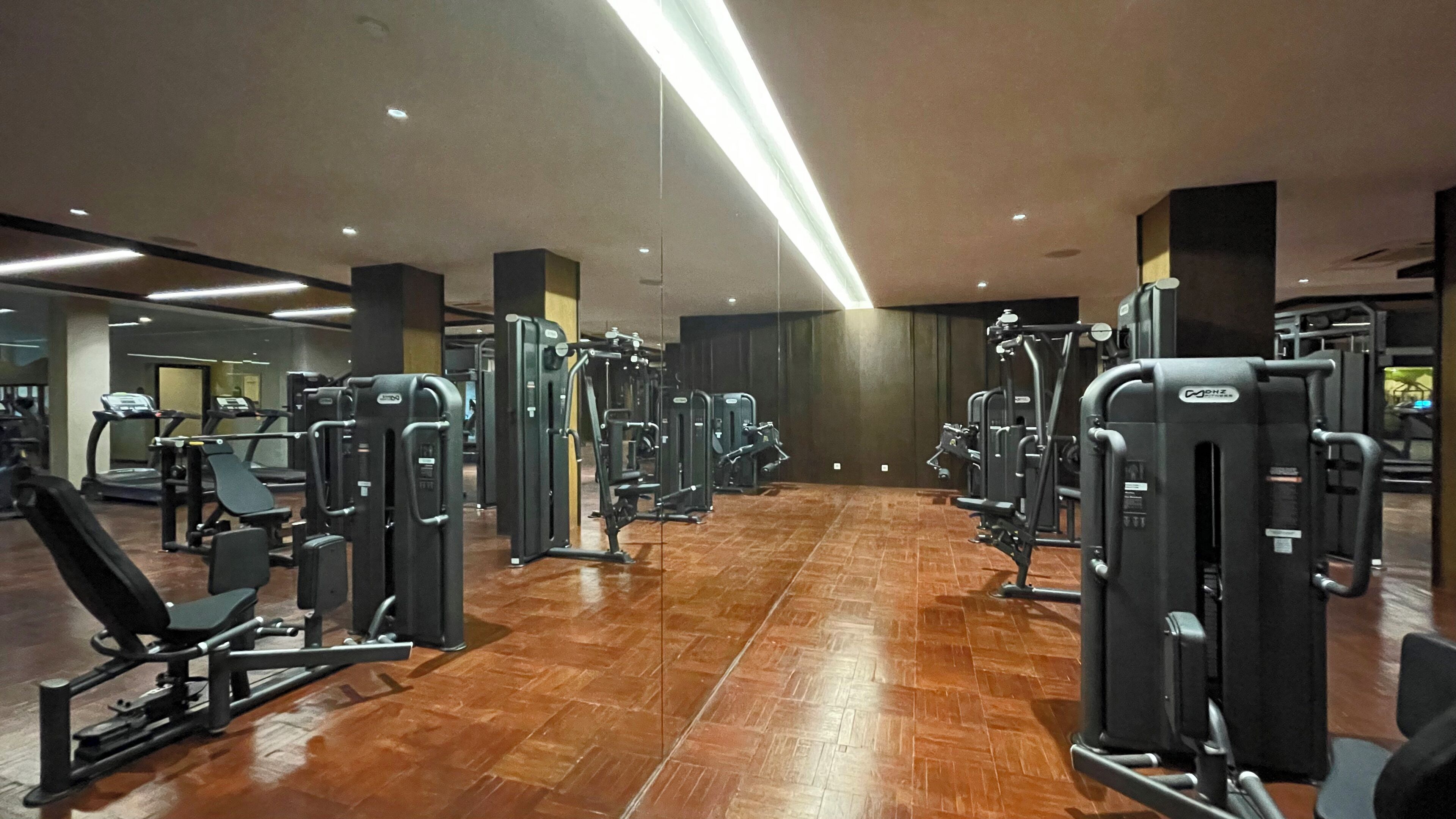 Academia com diversos aparelhos de exercícios, piso de madeira e design moderno no teto.