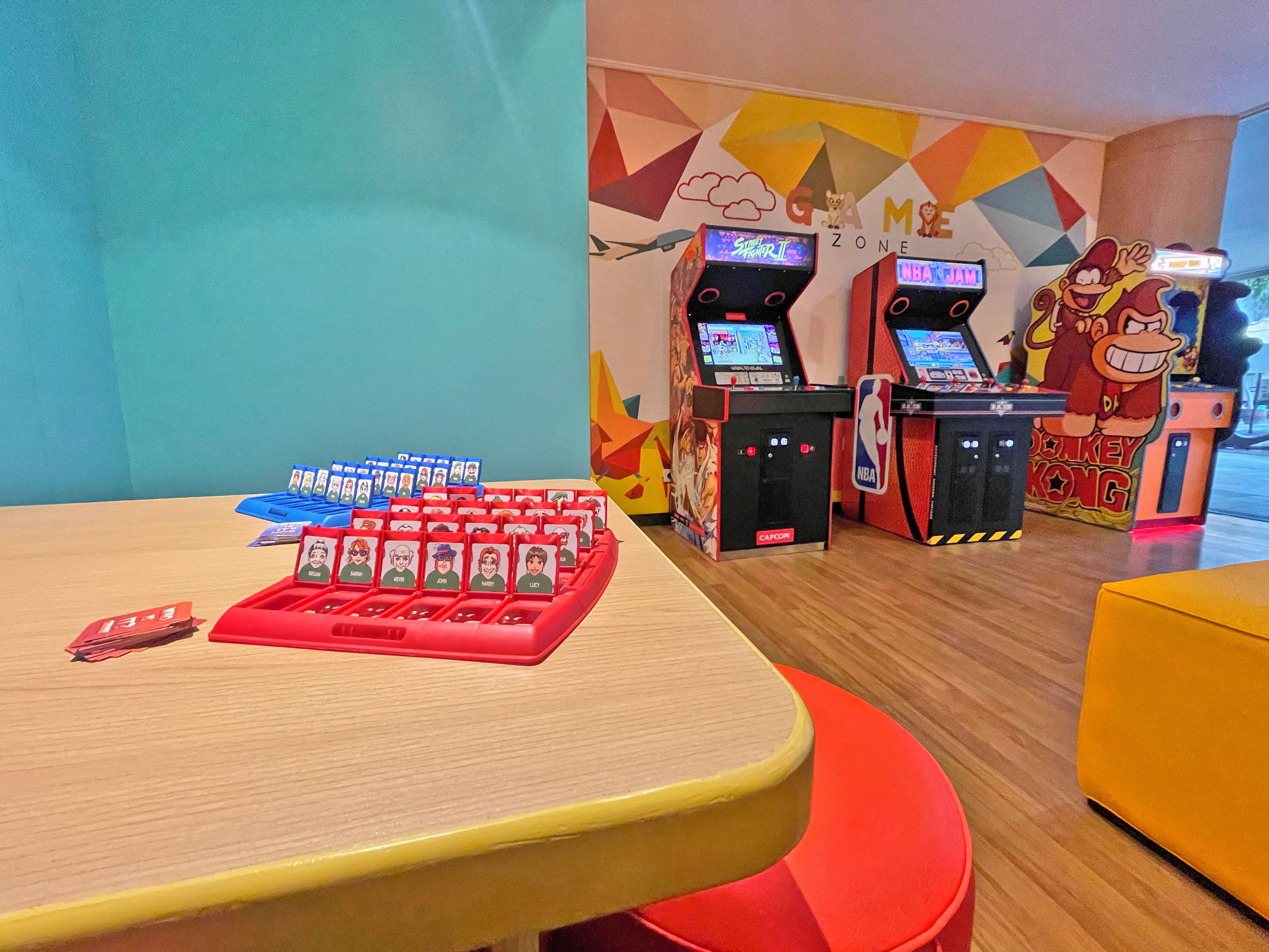 Un espace de jeux comprenant plusieurs bornes d’arcade, un espace dédié aux jeux de cartes et une fresque murale colorée.