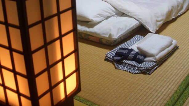 Una habitación japonesa tradicional con suelo de tatami, una cama con ropa blanca y un adorno rojo, una lámpara de pie de madera, y una mesita con ropa doblada y una toalla.