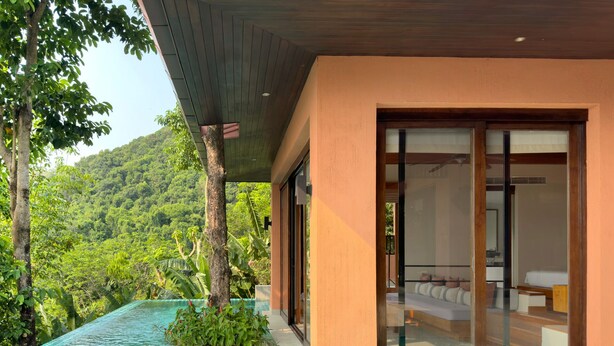 Una casa moderna con puerta de cristal que da a un dormitorio, rodeada de piscina y exuberante vegetación.