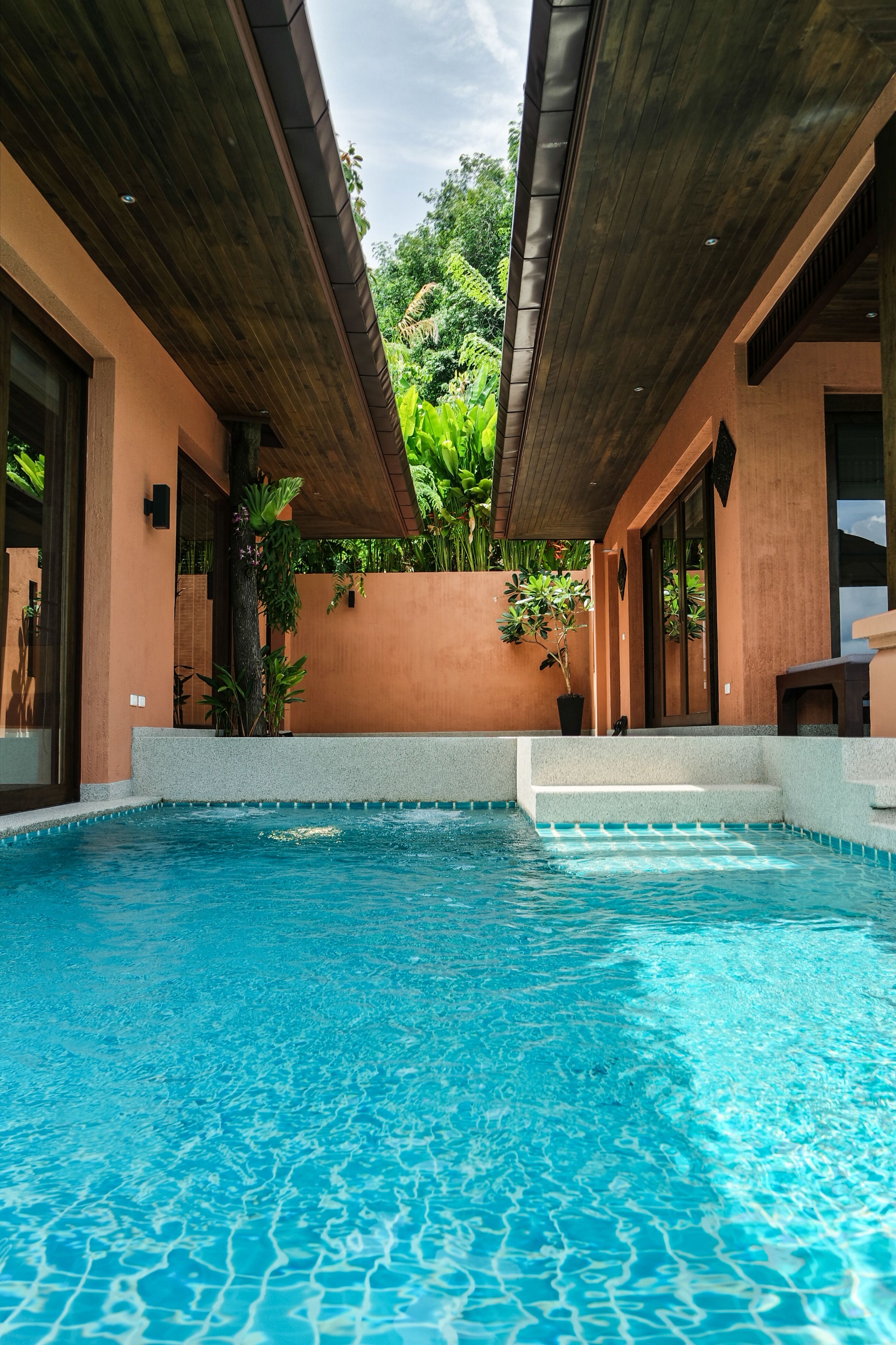A swimming pool with wooden beams overhead, surrounded by a peach-colored wall and lush greenery.