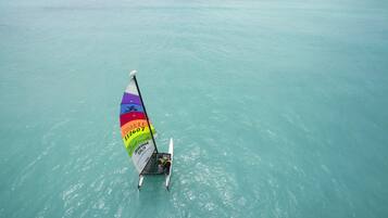 A windsurfing board with a colorful sail, a person standing on it, and clear turquoise water.