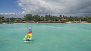 Un voilier sur une eau turquoise, avec un quai et des bungalows sur pilotis en arrière-plan.