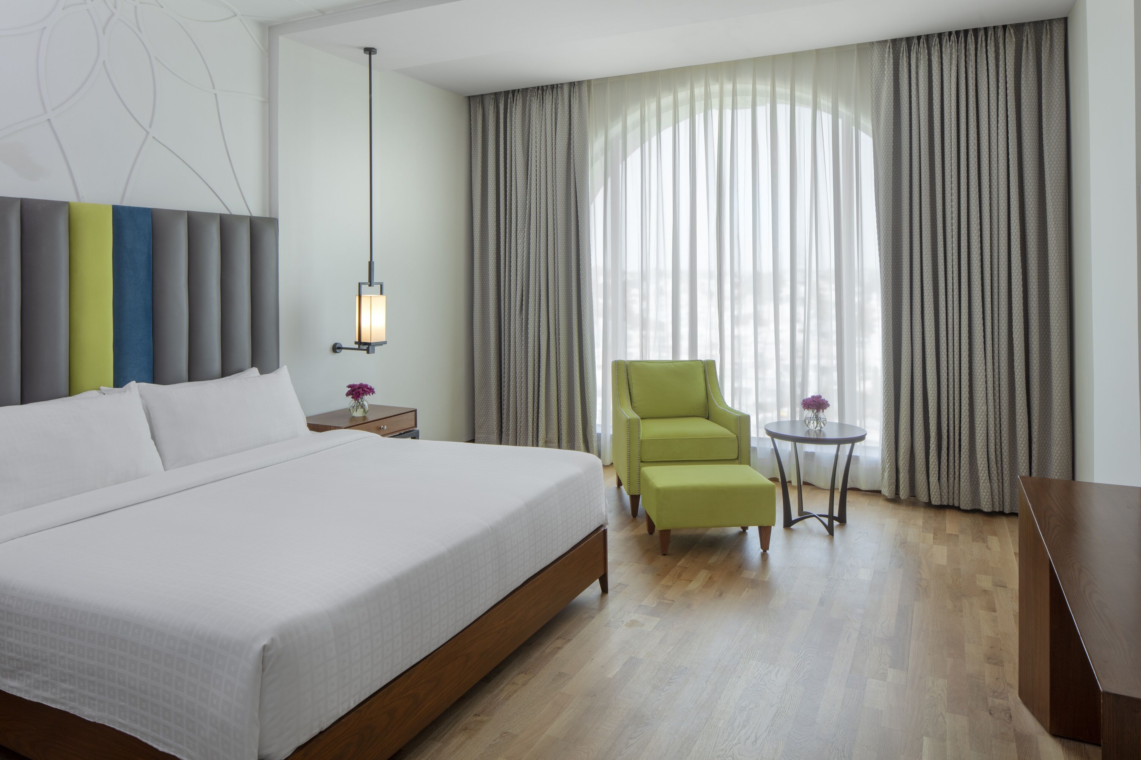 Habitación de hotel moderna con una cama grande, un sillón verde, una mesita con un florero y vistas a través de cortinas traslúcidas.