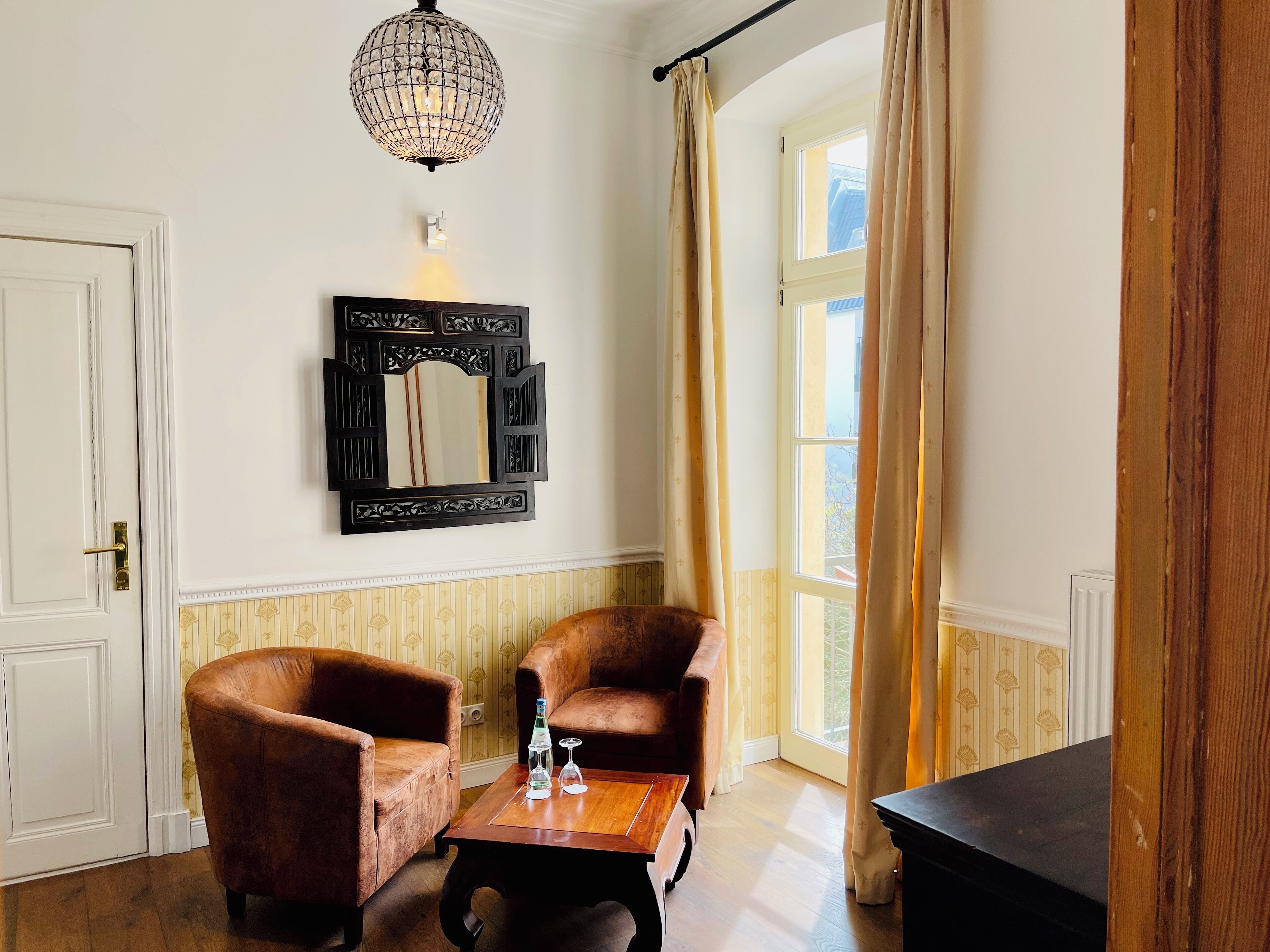 Une pièce comprenant deux fauteuils marron, une table basse en bois avec une bouteille et un verre, un miroir décoratif et un lustre.