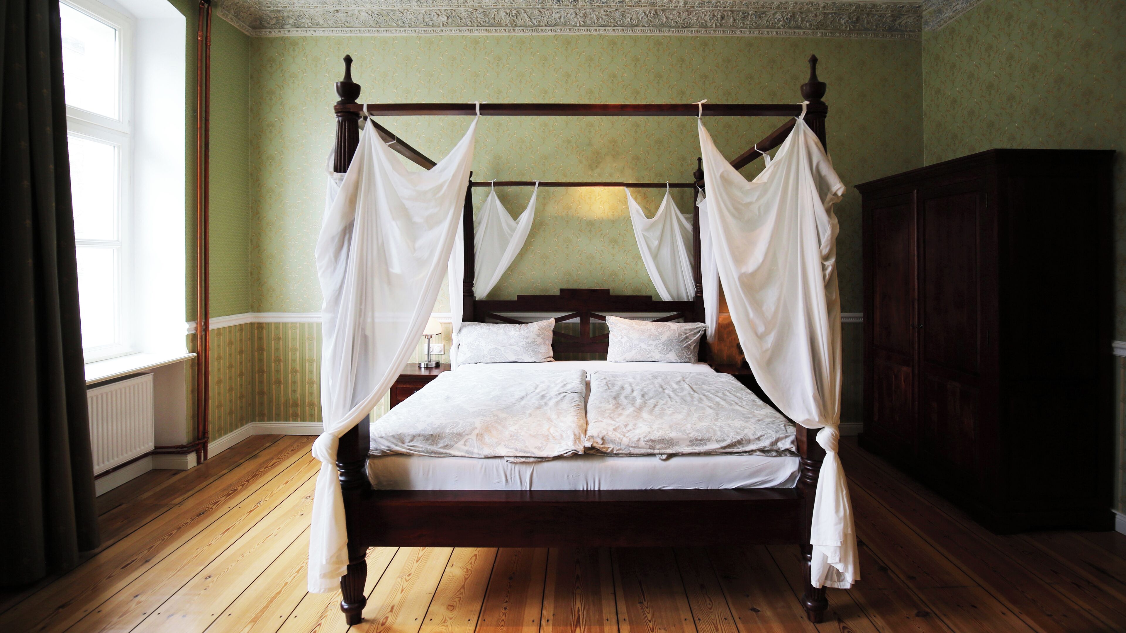 A four-poster canopy bed with white linens and a wooden frame, set in a room with green wallpaper and wooden flooring.