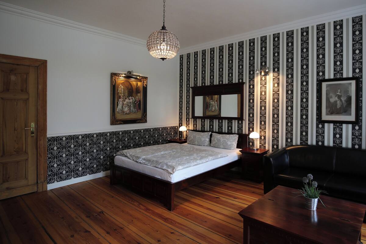 Un dormitorio con piso de madera, una cama con cabecera, un sofá de cuero negro, una mesita de noche de madera, un espejo montado en la pared y un candelabro.