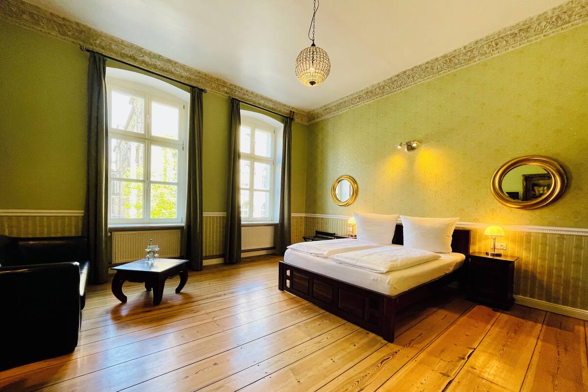 Una habitación de hotel con piso de madera, una cama con ropa blanca, un espejo redondo en la pared y una pequeña mesa con una lámpara.