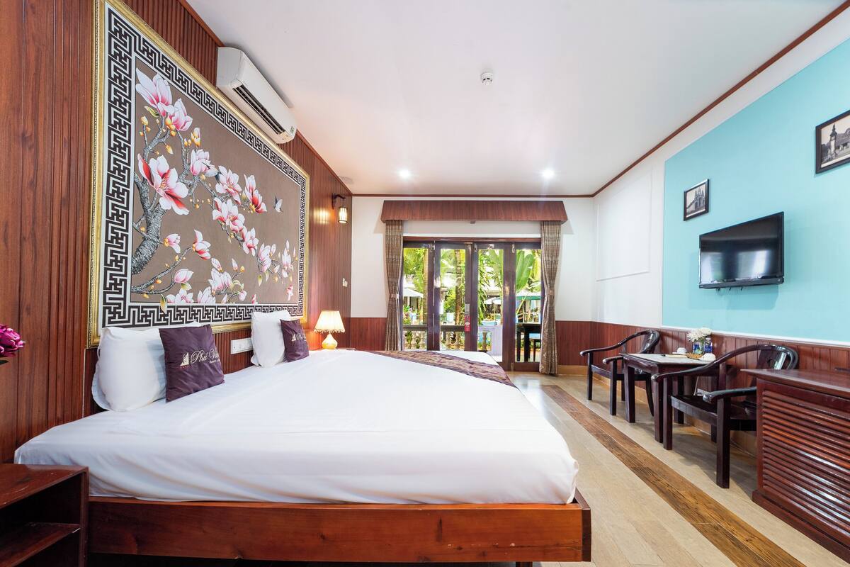 A hotel room with a large bed, wooden headboard, a TV mounted on the wall, and a desk with chairs.