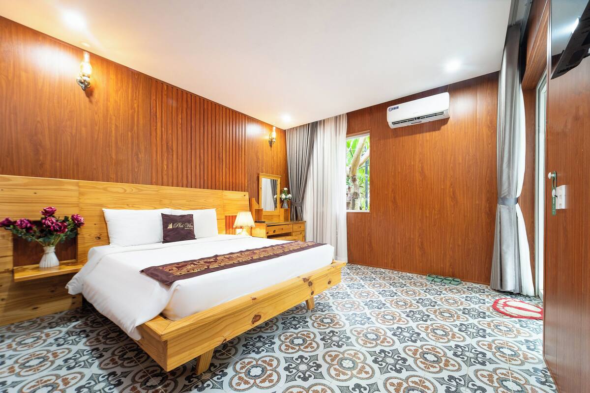 A hotel room with a wooden headboard, a large bed, a nightstand, a mirror, a window with curtains, and a patterned floor.