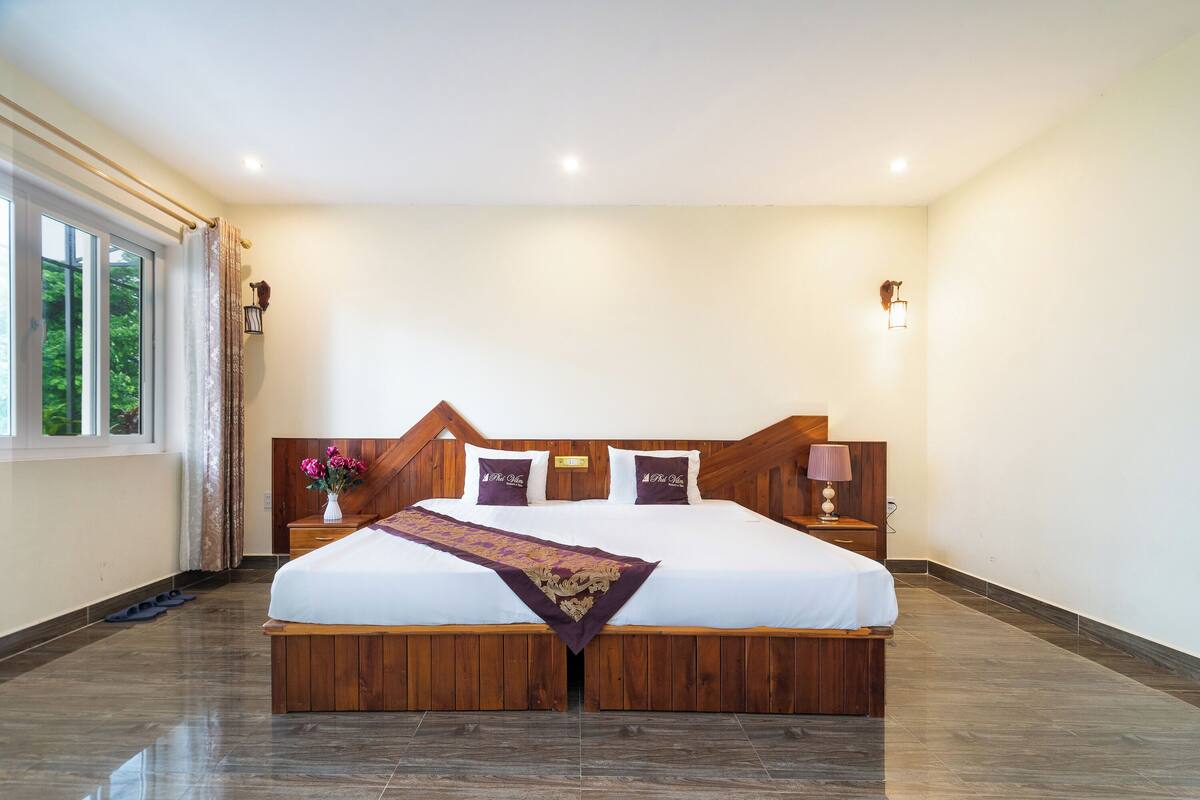 A neatly arranged bedroom with a large bed, wooden headboard, bedside lamps, and a window with curtains.