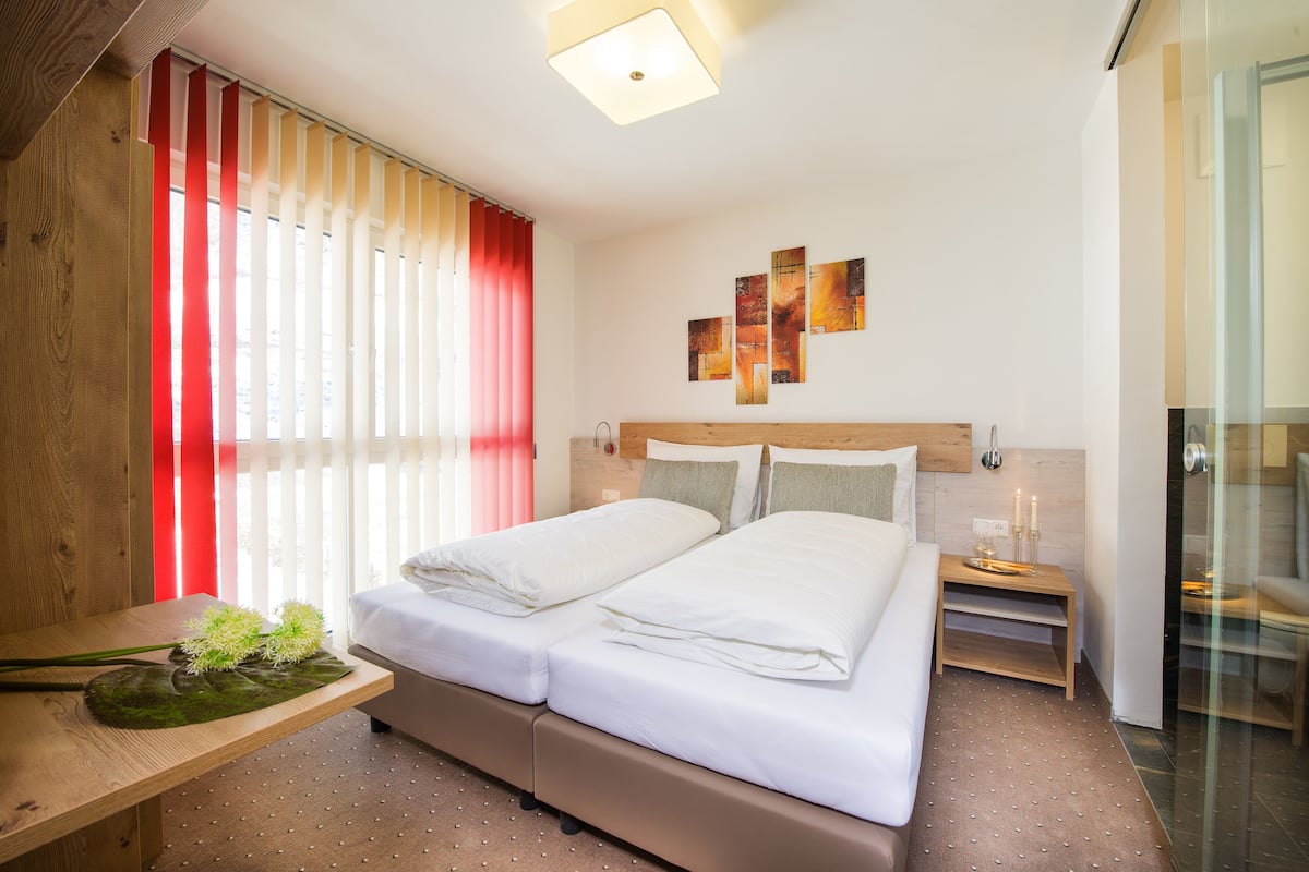 A modern hotel room with a large bed, a wooden nightstand, a small table with a plant, and a glass shower area.