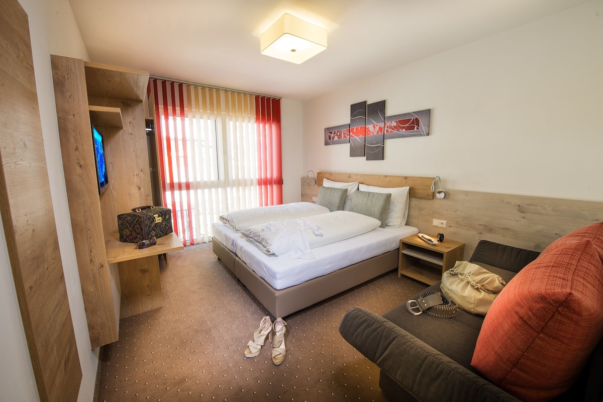A hotel room with a bed, a sofa, a TV, a wooden shelf, and a window with curtains.