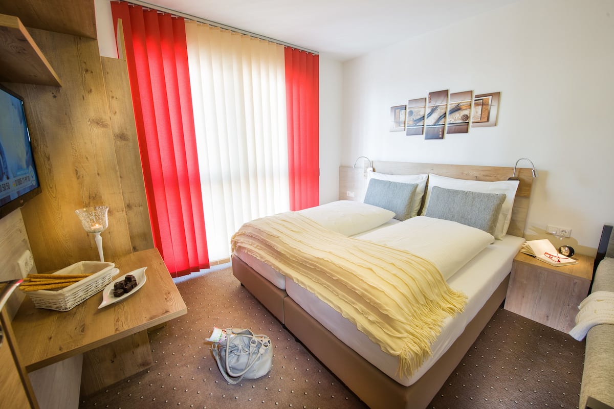 A hotel room with a bed, bedside tables, a TV, and a window with red curtains.