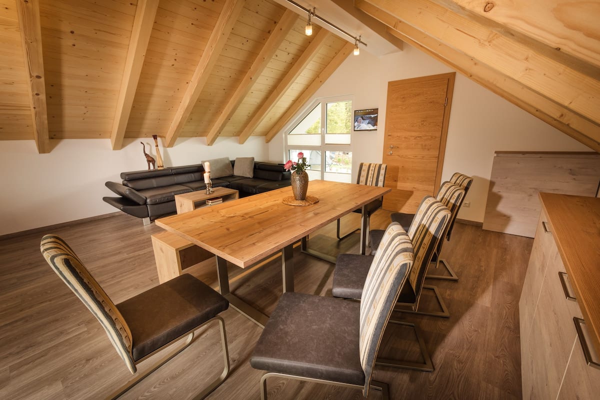 A dining area with a wooden table and chairs, a black leather sofa, and a wooden ceiling with exposed beams.