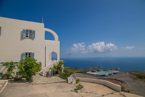 Un edificio blanco con contraventanas azules, un área de piscina con sombrillas y vista al mar.