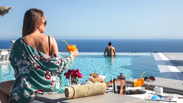 A person relaxing by a poolside table with a drink, a book, and a flower vase.