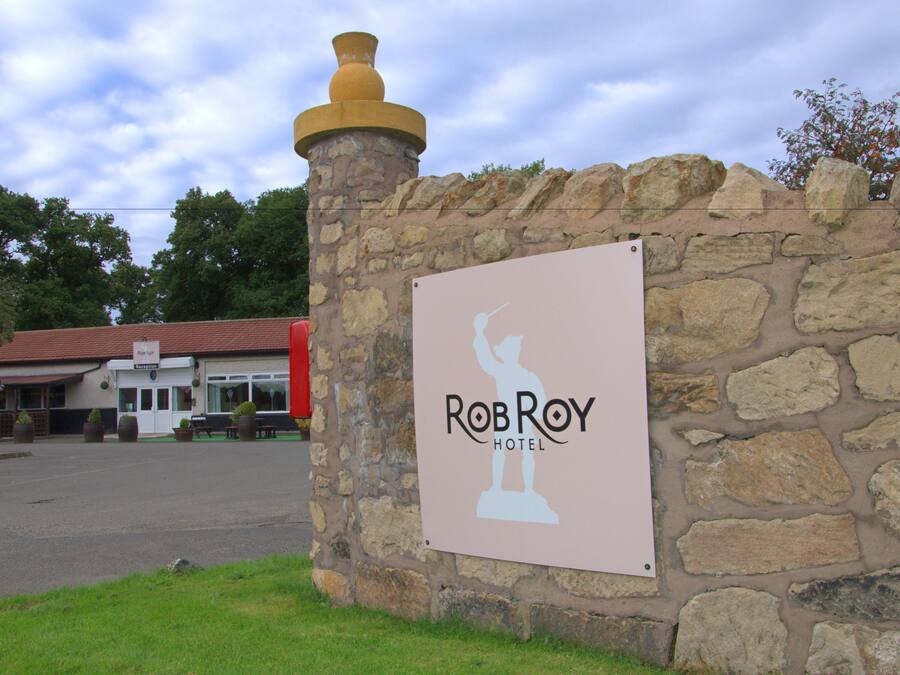 A sign for RobRoy Hotel with a stone wall and a building in the background.