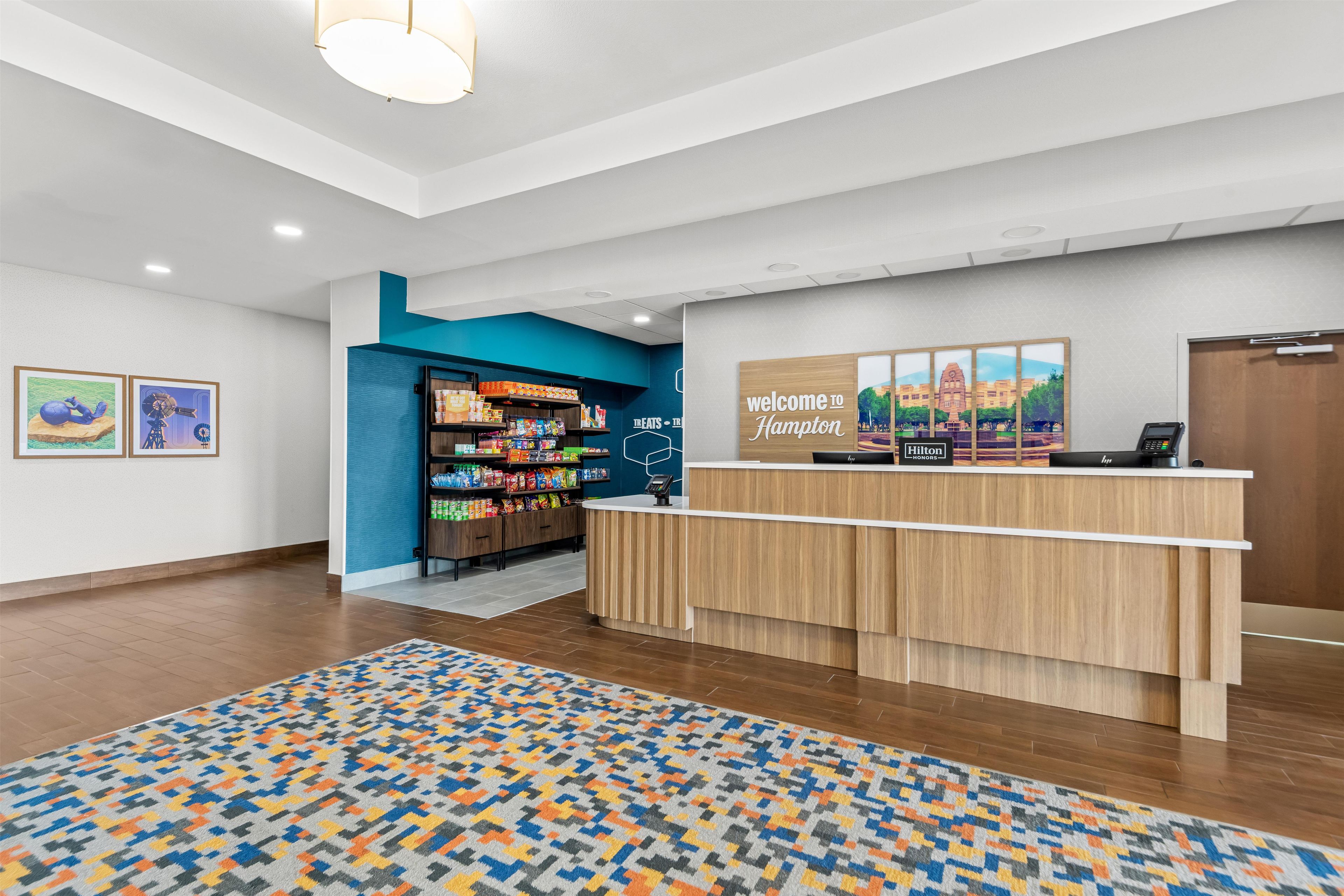 A hotel reception area with a wooden reception desk, a blue wall with a food display, and a 'Welcome to Hampton' sign.