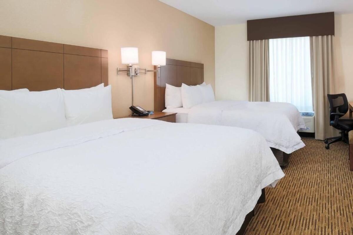Habitación de hotel con dos camas, un escritorio, una silla y una ventana con cortinas.