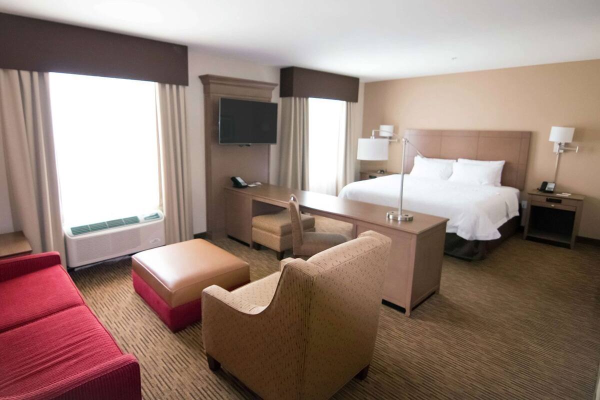 Habitación de hotel con cama, escritorio, televisión, sofá rojo, sillón color crema y ventana con cortinas.