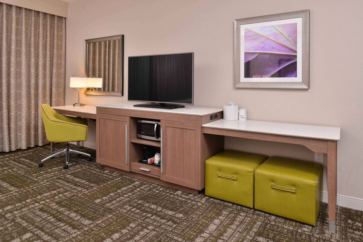 A hotel room with a desk, a television, a green chair, a microwave, and a small green storage unit.