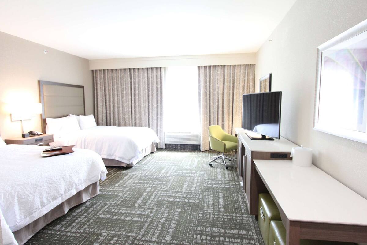 A hotel room with two beds, a desk, a chair, a television, and a large window with curtains.
