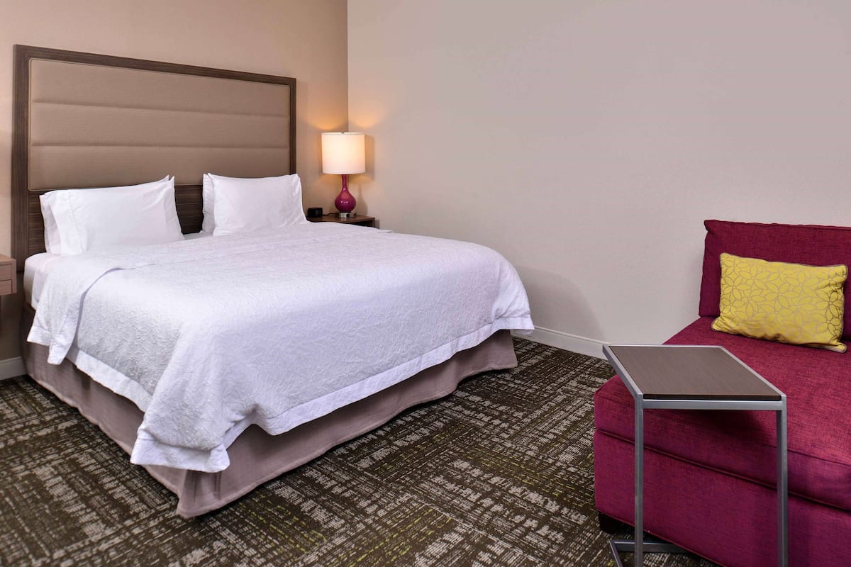 A neatly made bed with a headboard, a bedside lamp, a small table, and a red chair in a hotel room.