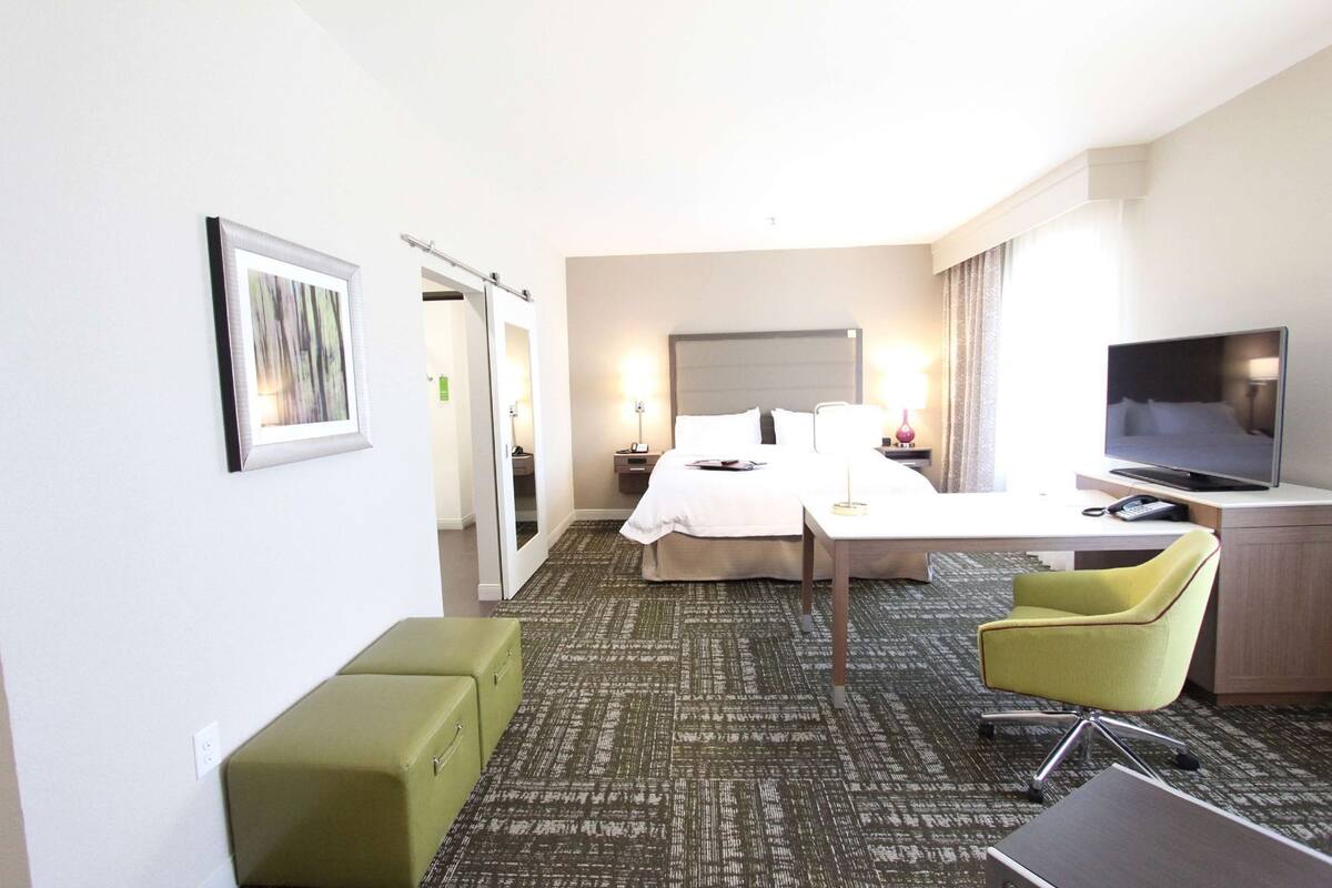 A hotel room with a bed, a desk, a TV, a chair, and a green ottoman.