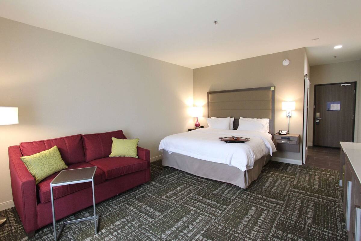 A hotel room with a bed, a red sofa, a small table, and a nightstand with a lamp.